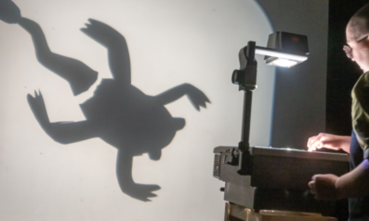 Madeline Shuron, wearing denim overalls and glasses, uses a vintage overhead projector to share the cut-out image of a frog losing its tail. We see the silhouette of this large-scale creature.