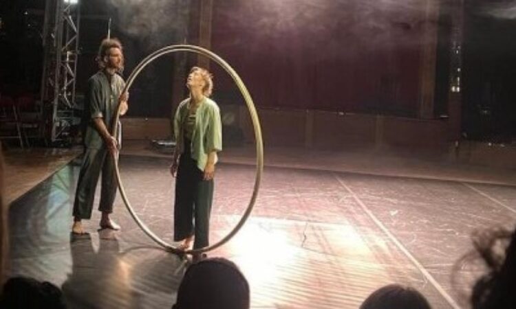 Hanschitz and Belerer, both in neutral green costumes, stand next to each other, Belerer holding the Cyr wheel while Hanschitz looks upwards to the top of the wheel. Hanschitz's foot rests on the bottom of the wheel as if about to mount it.