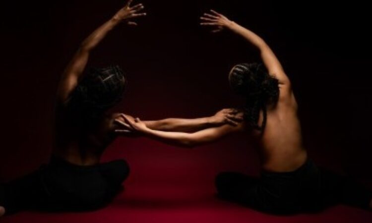 Two dancers sit on a red floor in front of a black backdrop with their hair braided into updos with cornrows. They are topless with their bare backs facing the camera, wearing black leggings. They mirror each other, leaning toward one another with one hand outstretched, resting on the other’s shoulder. The other arm arcs overhead, also in the direction of the other.