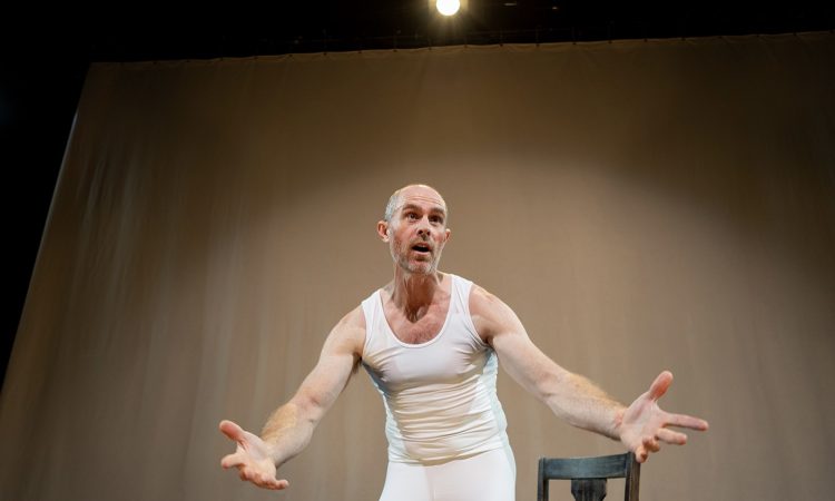 A bald white man wears a tight white tank top and white tights. He leans toward the audience, forehead wrinkled and arms outstretched in a questioning manner.