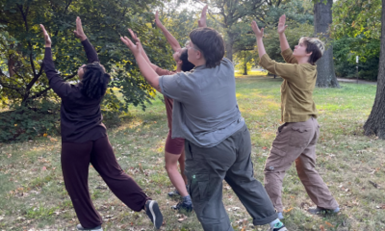 a group of four individuals stand together with arms lifted overhead making a clapping gesture with their hands. They are outside in the grass, surrounded by trees and other greenery. Their outfits are earth tones - browns, greens, and shades of grey.
