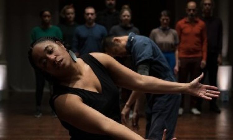 Shavon Norris stands in front of an audience wearing black clothing, large circular earrings, and a few finger rings. She is tilted to one side, facing the camera with eyes closed, arms outstretched in front of her, and palms facing upward.