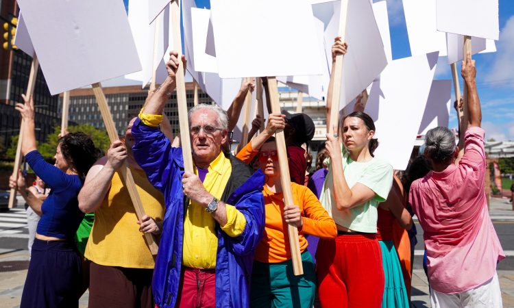 A line of people in bright, solid colored clothes walk across Dilworth Park. From this camera angle, we catch the sides of blank white signs they hold up, and see three to eight feet of space between each marcher. They pass a grid of sprouting water fountains in the foreground and the background. Behind them, large downtown buildings, both modern metal and glass, and old stone, loom.