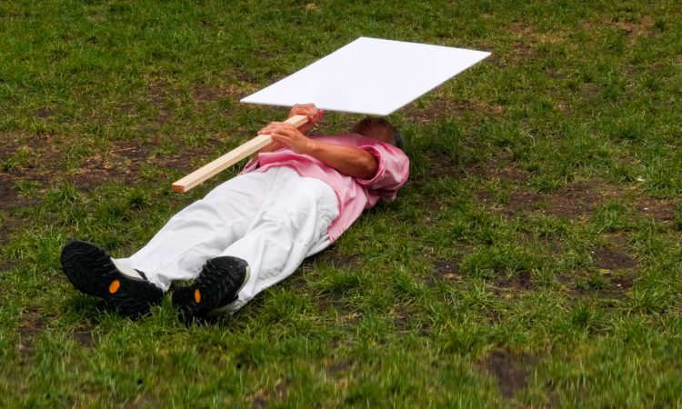 A man in bladk shoes, white pants, and a pink button up shirt lies on the ground on green grass. He holds a blank sign on a wooden pole horizontally over his body
