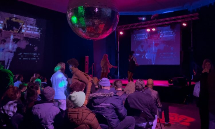 The audience is awash in purple; the room is dim and the discoball floats overhead, reflecting green and red and purple. There is projection on the back wall of the stage that is purple with blurry outlines of people, and another projection on a sidewall with people dressed in white in a dress and clownish costume. The audience mills about, some with drinks in hand. A performer in a black dress sings into a microphone onstage while someone else appears to be facing them, speaking to them.