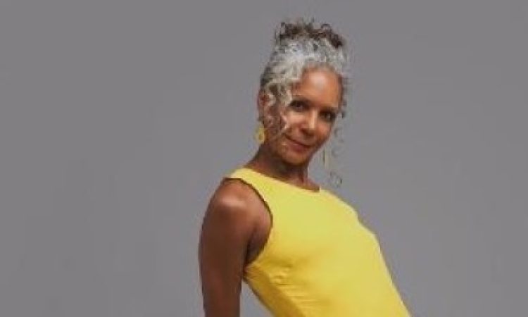 Jennifer Jones, a middle-aged African American woman stands, left hip jutting out. She wears a bright, bellowing yellow dress, and her curly gray hair frames her face.