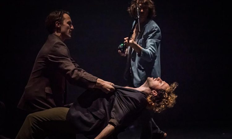 One dancer grabs the other by the collar, suspending him in midair. The lifted dancer’s head tilts back and his chest opens, embodying a forced exposure and a sense of loss of control. The two dancers appear to have just been in a struggle. Behind them, a performer holding a microphone stands in the light, as if witnessing or intervening in this moment.