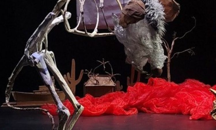A large sculpture of a muskrat stands in a blackbox theatre. The muskrat is mostly skeleton with a furry head and ears. There is some red fabric on the floor in the background and a ball of red wiring sitting beneath the muskrat.