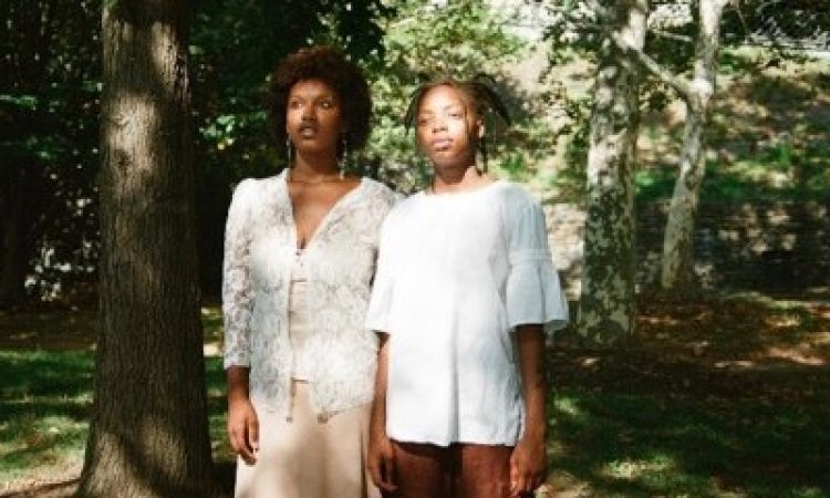 Two Black performers stand side-by-side, wearing pedestrian clothing – lacy, patterned white tops layered over shades of beige and brown. Their gaze extends out to their right, converging on an unknown entity in the distance. Surrounding them, a park with green grass and trees foregrounding urbanity.