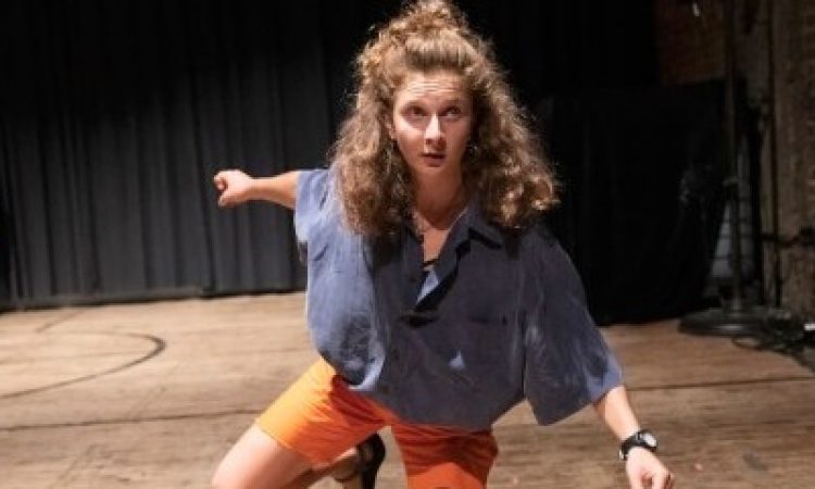 A dancer in a blue denim button down shirt and orange shorts is on one knee with her other leg raised and bent slightly behind her. She has long curly hair and peers out into the audience with her arms outstretched.