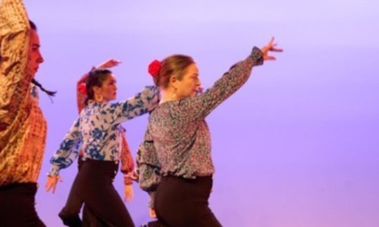 A group of women in printed blouses are moving across the stage. The two women in front have one arm raised on the forward diagonal. 