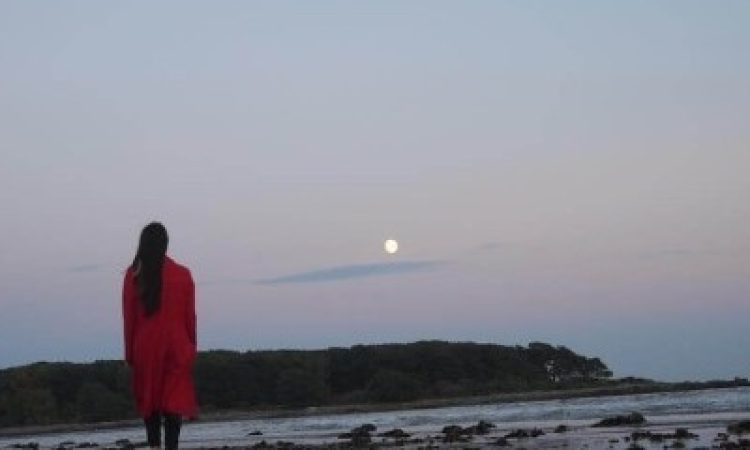 View of Nichole Canuso from behind standing on the beach, her left foot in mid-footfall, at night in an orange coat with the full moon high in the sky.