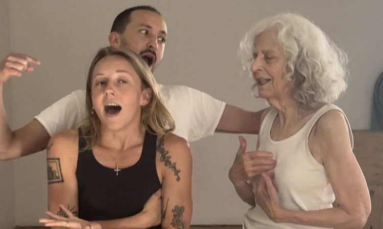 three performers wearing plain white or black tanks/tees are mid gesture as if having a family argument. The performer in the front hunches their shoulders, crosses their arms, and has mouth open, perhaps in the middle of a sentence. Behind them, another performer looks to be yelling and pointing at the third performer, a hand on their shoulder, who has eyes closed, a soft smile, and holds their fingertips to the center of their chest.