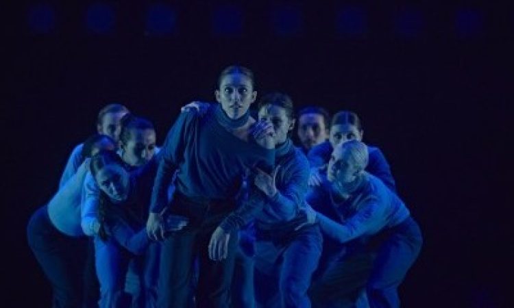 In blue lighting, a crowd of dancers hover over one another looking forward into the camera lens. One dancer stands in the forwardmost position with the group grasping their shoulders, arms, and legs, as if to constrain their forward motion.