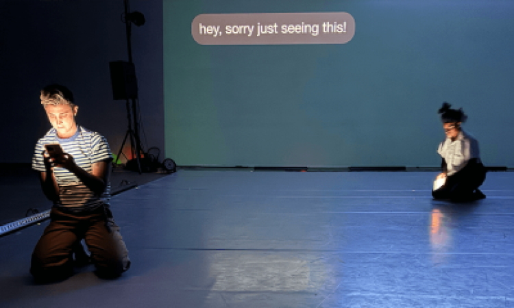 Two dancers kneel on the ground, illuminated by their phone screens. Behind them a projected text bubble reads "hey, sorry just seeing this!"