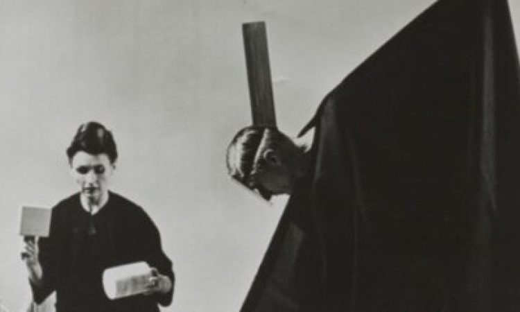 A black and white photo of Lucia Dlugoszewski kneeling on the floor holding a cylinder and a box shaped item in either hand. in front of her sits a pile of wooden objects and instruments. beside her, Hawkins, draped in a black cape-like garment which reveals only his head, tilts toward Dlugoszewski and creates a triangular shape with the garment.