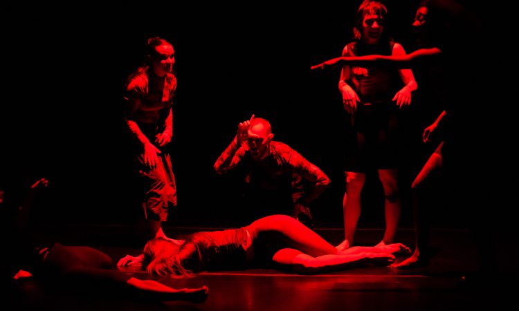 The light’s red, the stage black. Dramatic. Devilish. Contrast and shadow are high on the six people in frame. Dead center, a dancer lays. Around her, four people point, laugh, and crouch over her body making the “loser” L on their forehead. In the bottom left corner, someone's leg, hip, and reaching hand are just in frame.