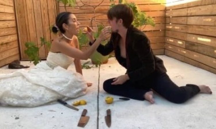 A bride and groom sit on white concrete barefoot with knives and lemons in front of them, feeding each other lemon wedges with a fig tree in a pot and wood fencing in the background.