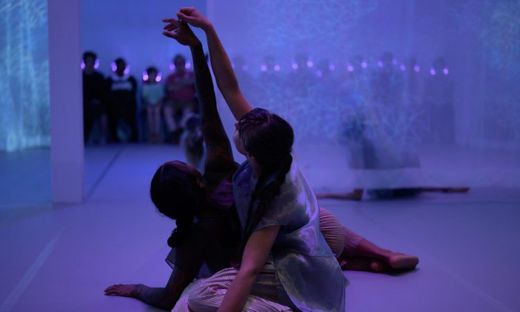 Chloe Marie Newton, a black female dancer wearing a gauzy dark blue tank and Helen Hale, a white female dancer with a long brown ponytail wearing shimmery silver, sit on a light grey marley floor, reclined on one hip with knees gently stacked and one arm intertwined with the other, reaching upwards. They have their backs to us and around and beyond them, we can see pale, cool-toned, sheer fabric. Through and beyond the fabric, we see a row of shadowy-figured audience members, seated and wearing headphones that emit a blue glow.
