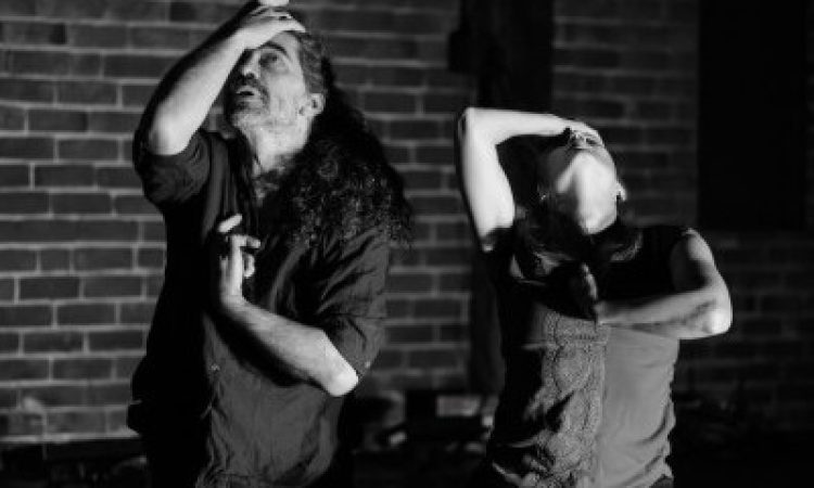 A black-and-white photo of two dancers in a brick-walled room. One, masc-presenting, has long curly hair and peeks out at the ceiling, mouth slightly open in expressive thought, one hand bent to touch their forehead, shielding half of their face. The other hand rests against the center of their body. A second dancer stands to their left, mirroring this pose with face tilted all the way to the sky and taut arms.
