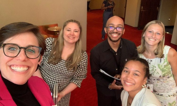 A group of five individuals, dressed in business attire, all gather together for a selfie in the velvet-carpeted lobby of the John F. Kennedy Center for the Performing Arts. Jane, at the front left, is a white, brunette woman with a medium pixie cut. Clad in a magenta blazer and black turtleneck, Jane dons a bright, bespectacled smile. Grinning behind Jane, Mallory, a white woman with dirty blonde hair, wears a black and white gingham dress and holds a silver clasp. Malik, a tawny-skinned Black man in a black button-down and trousers, stands beaming at Mallory’s left. Allison and Chloe, dressed in a white button-down and a floral dress respectively, lean into the photo, offering their smiles as well.