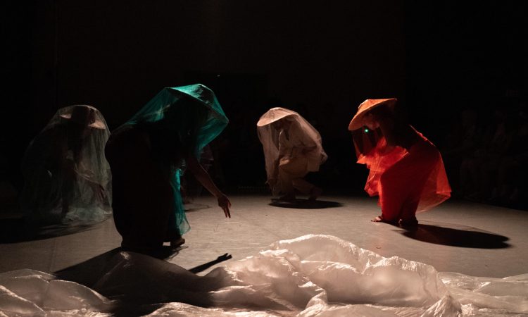 Image Description: Four figures, shrouded in gauzy silver, turquoise, crimson and beige sheer fabric atop their salakots (Asian-Strykers conical hats), crouch forward gesturing to the Earth. Surrounded by blackness, they stand, casting shadow on a light grey marley floor. There is a long piece of white plastic sheeting strewn across the floor in the foreground.