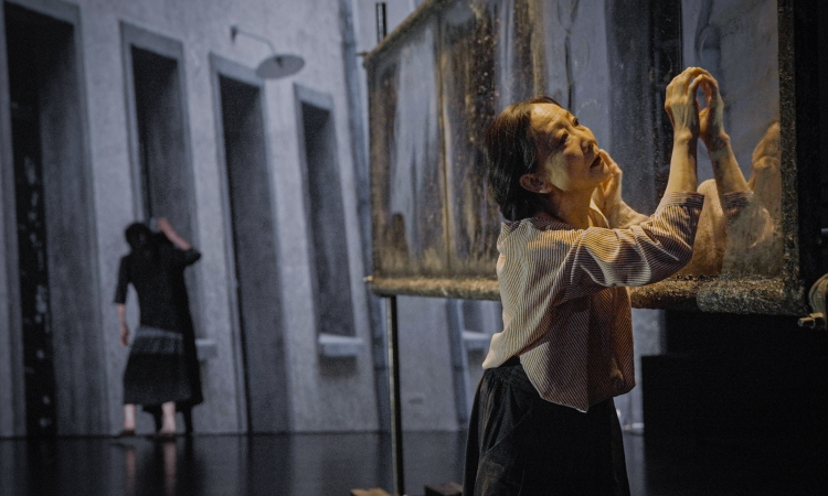 A Chinese woman, Wen Hui, stands in a grey, concrete room with three long vertical doorways. A figure dressed in black, whose back is turned to the camera, in the background. She gently touches an old, heavy, rust-encrusted horizontal mirror, dusty from time, hanging in the air. Warm light shines on her face, a worn down curiosity in her expression. 